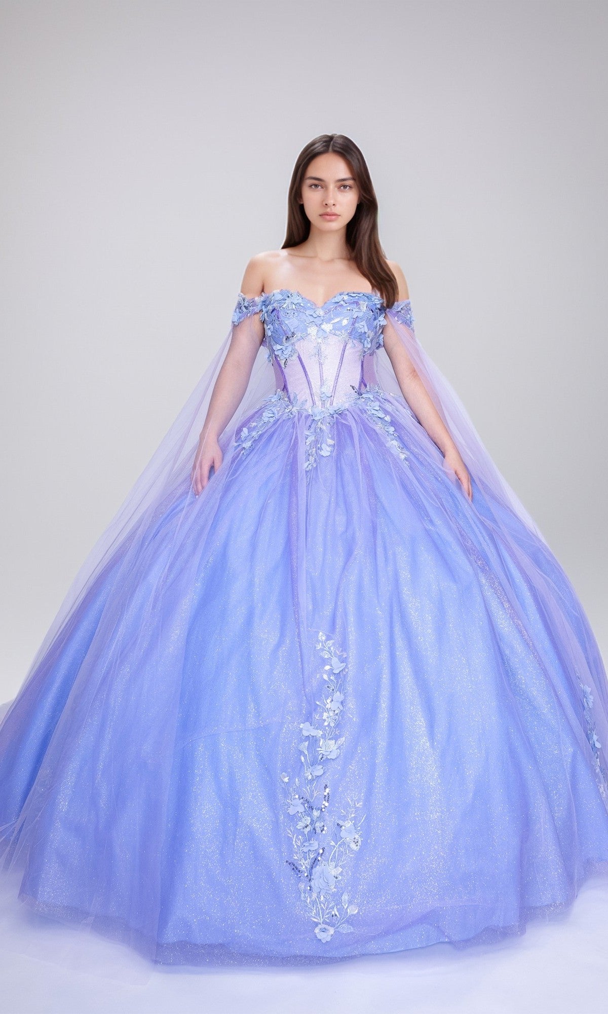 A woman wears the Quinceanera Dress Q117 by Nina Canacci in lilac/baby blue, featuring off-the-shoulder sleeves, floral appliqués, and a glitter tulle skirt, set against a plain light background.